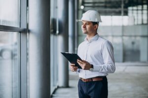 Engenheiro de negócios, usando um capacete de segurança branco e camisa social, segurando uma prancheta enquanto observa atentamente o interior de um prédio em construção, através de uma janela de vidro.