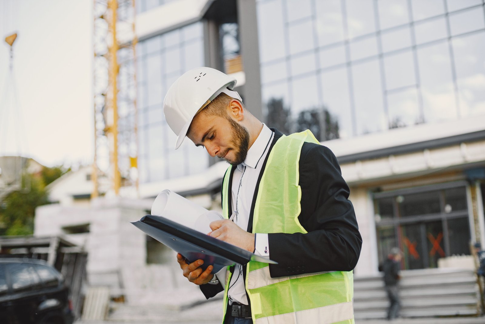 A imagem mostra um jovem engenheiro, vestido com um capacete de segurança branco e um colete refletivo verde, analisando atentamente um conjunto de projetos em frente a um edifício em construção. Ele segura uma prancheta e parece concentrado nos detalhes do documento, com o canteiro de obras e uma estrutura de vidro ao fundo.