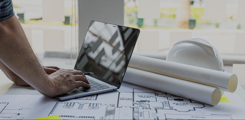 A imagem mostra uma cena típica de trabalho de engenharia. Ao centro, um homem está utilizando um laptop sobre uma mesa repleta de plantas de projeto arquitetônico enroladas. Ao fundo, são visíveis algumas estruturas de uma construção e uma janela com luz natural.