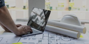 A imagem mostra uma cena típica de trabalho de engenharia. Ao centro, um homem está utilizando um laptop sobre uma mesa repleta de plantas de projeto arquitetônico enroladas. Ao fundo, são visíveis algumas estruturas de uma construção e uma janela com luz natural.