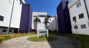 Imagem do prédio industrial da Sanofi, sob um céu claro. O prédio é amplo e possui paredes predominantemente brancas, com destaques em roxo escuro. Há um pequeno jardim bem cuidado na frente, com gramado verde e algumas palmeiras. Uma placa com o logo da Sanofi está visível na frente do prédio.