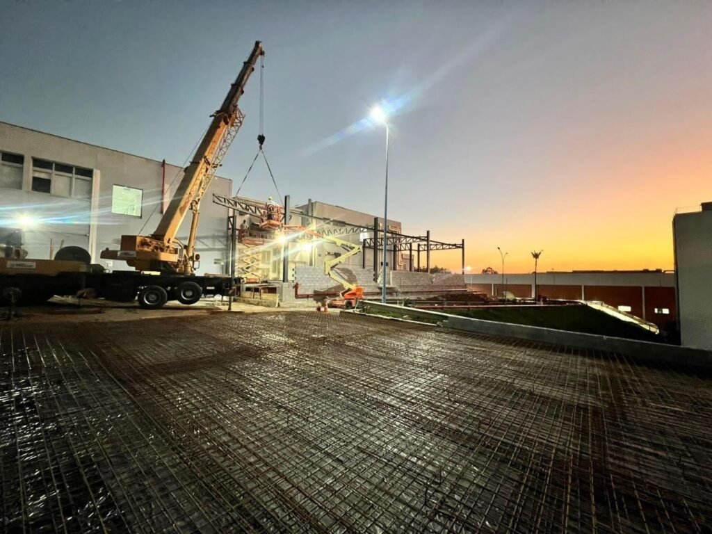 Imagem de uma obra de construção ao entardecer. Uma grande grua amarela está em operação, erguendo materiais de construção.