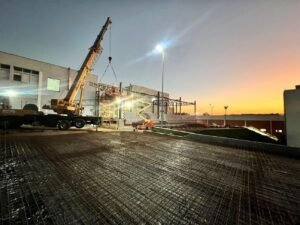 Imagem de uma obra de construção ao entardecer. Uma grande grua amarela está em operação, erguendo materiais de construção.