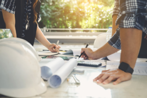 A imagem exibe uma reunião de equipe ao ar livre focada em engenharia. Três profissionais, dois homens e uma mulher, estão sentados em torno de uma mesa com documentos e planos de construção. A mesa também contém um capacete de segurança, um notebook, e canetas. Eles discutem ativamente, rodeados por um ambiente verde e ensolarado.