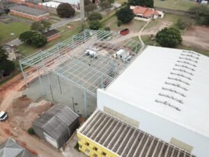 Foto aérea de uma construção industrial com estrutura metálica azulada em progresso. À esquerda, um terreno com equipamentos de obra e um galpão industrial adjacente com telhado branco.