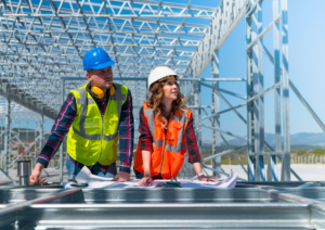 Dois engenheiros analisam plantas de um projeto em uma estrutura industrial em construção. Ambos estão equipados com EPIs, incluindo capacetes de segurança e coletes reflexivos, demonstrando o compromisso da Mabex com a segurança e a qualidade em obras de alta complexidade.